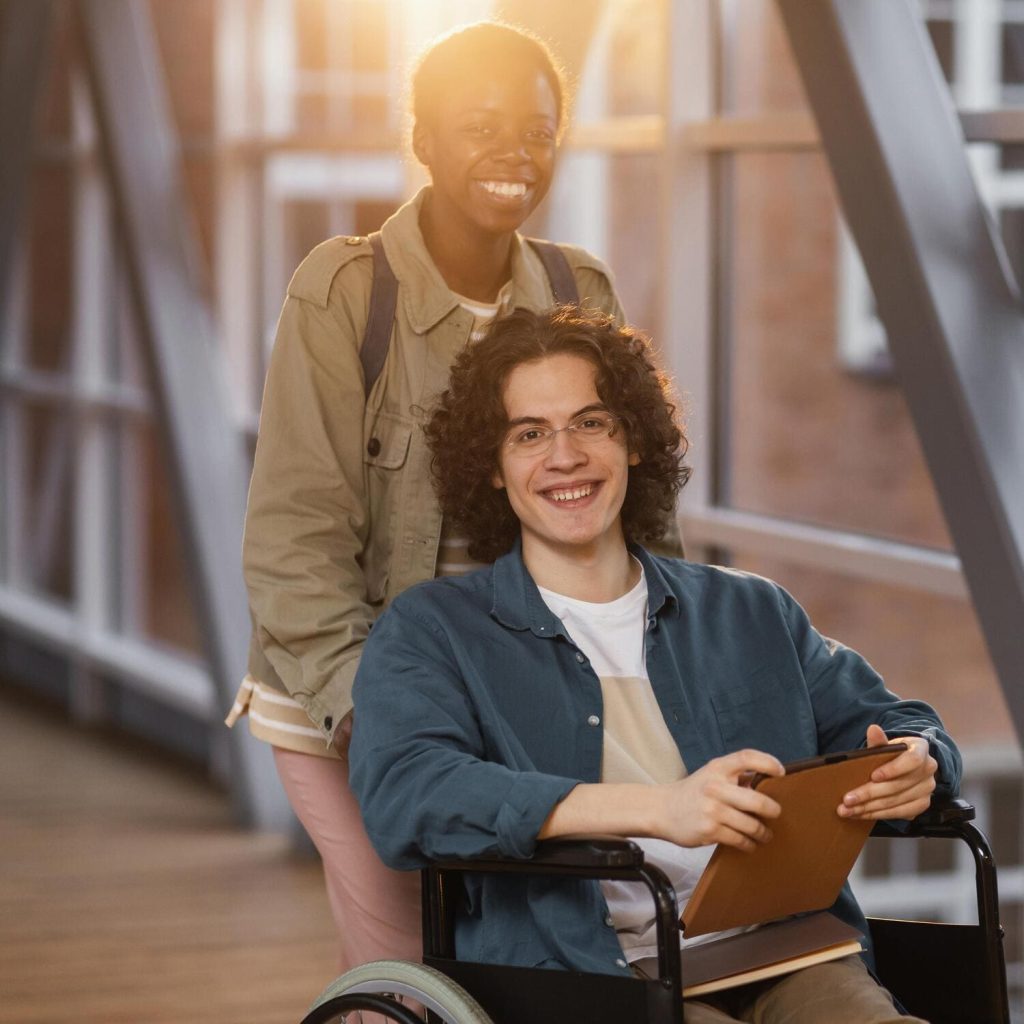 joven sonriente ayudando a su compañero en silla de ruedas mientras miran a la cámara